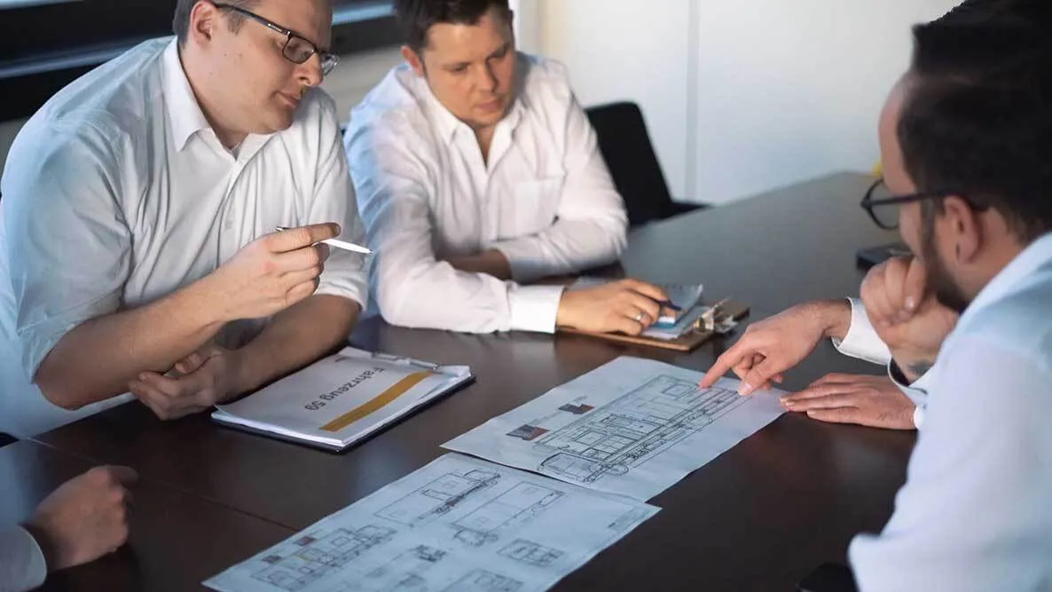 Drei Herren sitzen an einem Tisch und besprechen die geplante Inneneinrichtung, die als Zeichnungen auf dem Tisch liegen.